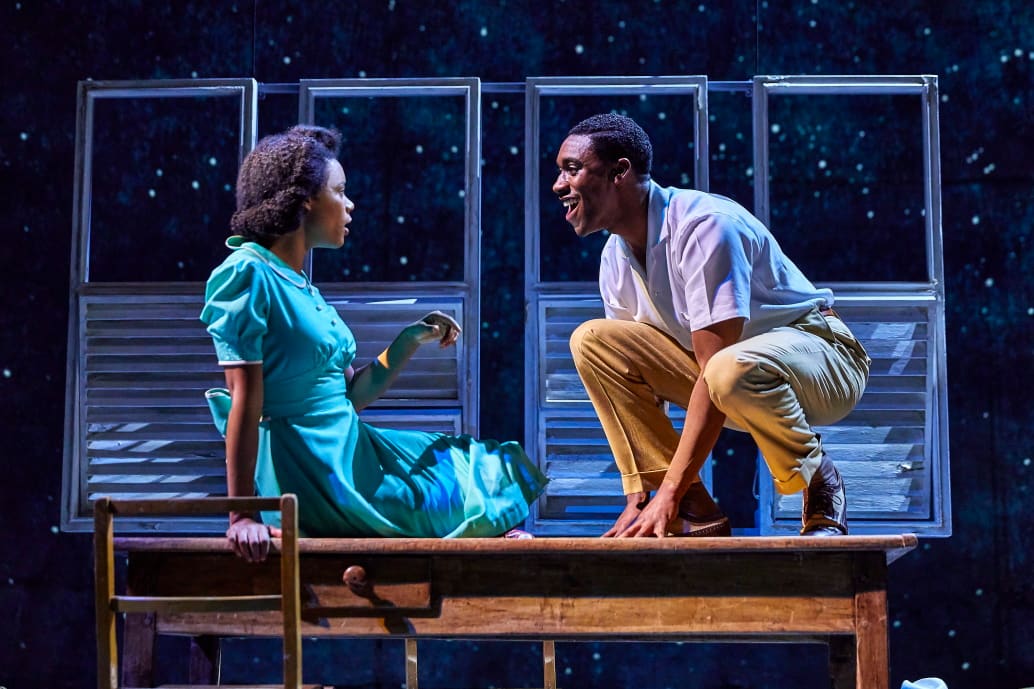 A production of Small Island featuring CJ Beckford and Leah Harvey who are sat on stage, facing each other and talking. Credie line: Small Island Photo by Brinkhoff Mögenburg